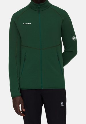 Mann trägt eine dunkelgrüne Jacke mit durchgehendem Reißverschluss und Mammut-Logo auf Brust und Ärmel, kombiniert mit schwarzen Hosen mit Mammut-Logo-Patch.