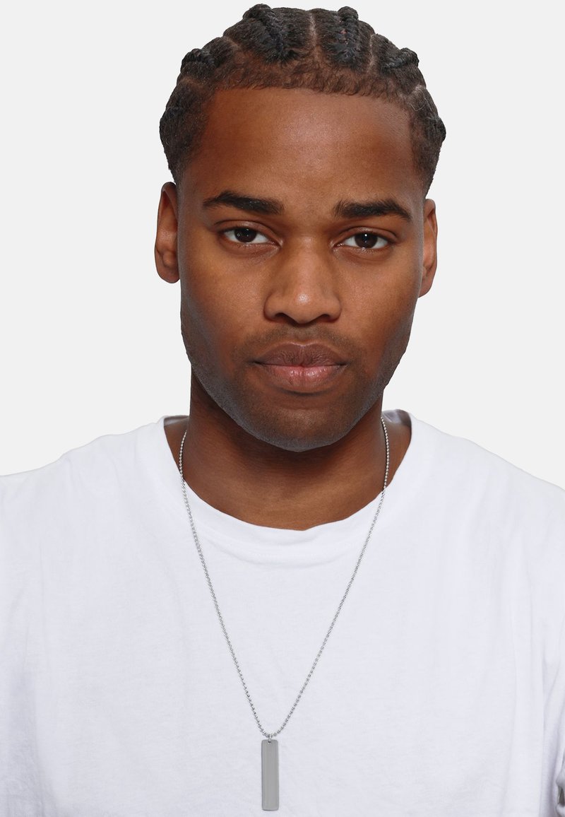 Collana con pendente d'argento su una catena sottile, indossata sopra una t-shirt bianca semplice. Capelli corti e raccolti in treccia. Pelle chiara, espressione neutra.