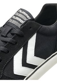 Schwarze Sneaker mit einem strukturierten Stoff-Obermaterial, weißen Lederakzenten in Form von Chevrons und einer schwarzen Zunge, die das "hummel"-Logo in Weiß zeigt.