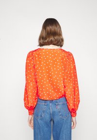 Femme aux cheveux bruns mi-longs portant un chemisier à manches longues orange à pois blancs et un jean taille haute bleu, vue de dos.
