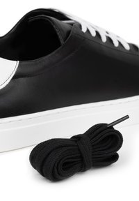 Czarne skórzane sneakersy z białymi akcentami, płaska biała gumowa podeszwa oraz czarne płaskie sznurowadła umieszczone obok buta. Gładka faktura.