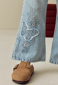 Ljusblå denimbyxor med utsvängd benpassform, dekorerade med fjäril- och blommönster i silvertråd; matchade med bruna mockaskor med rem och spänne.
