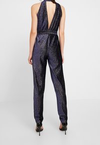 Mono azul marino con diseño de cuello halter, espalda descubierta y textura brillante. Cintura ajustada con piernas rectas, combinado con tacones altos.