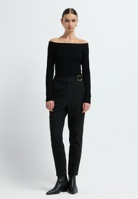 Vrouw die rechtop staat, gekleed in een zwart off-shoulder ribbeltop, zwarte high-waist broeken met een gouden gesp en zwarte enkelboots.