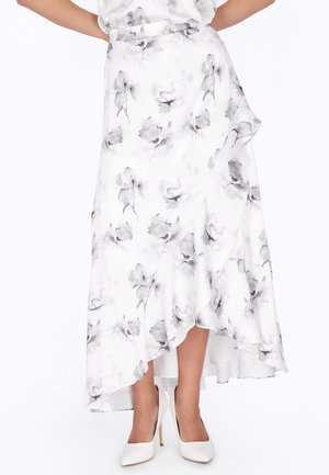 Jupe blanche à ourlet asymétrique avec imprimé floral gris, portée avec des escarpins blancs, les mains du modèle sur les hanches, sur un fond uni.