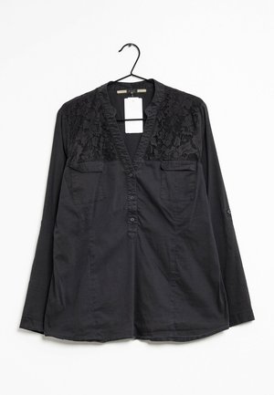 Blouse noire à manches longues avec des détails en dentelle sur les épaules, deux poches poitrine, col en V boutonné, suspendue à un cintre noir avec une étiquette blanche.