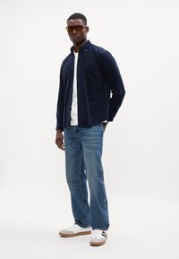 Marineblå bomulds skjorte med knapper, hvid T-shirt under, lyseblå jeans og hvide sneakers med gummisåle. Brune solbriller bæres.