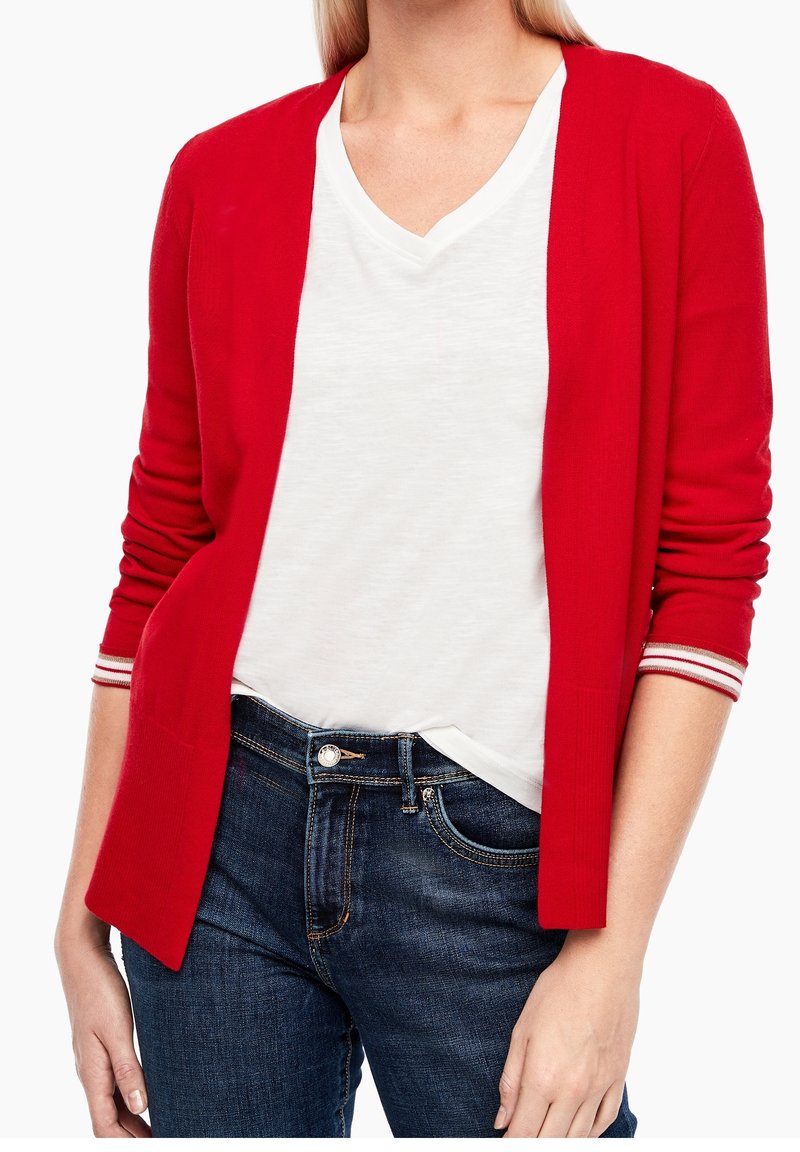 Femme portant un cardigan rouge avec des poignets rayés sur une chemise blanche à col en V et un jean bleu denim, debout devant un fond blanc.