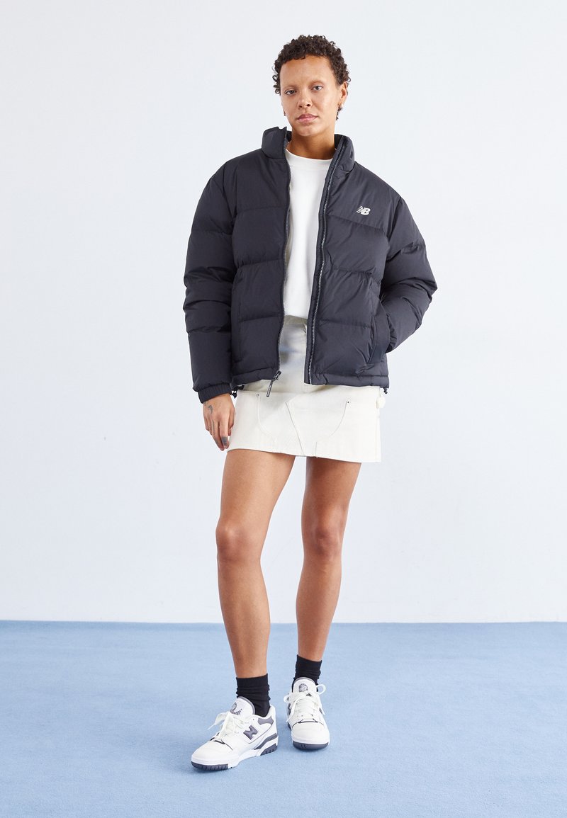 Schwarze Daunenjacke mit Reißverschluss und hohem Kragen, ausgestattet mit einem Logo auf der Brust. Getragen über einem weißen Oberteil und kombiniert mit einem beigen Rock.