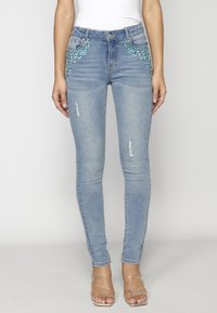 Jeans de mezclilla azul ajustados con adornos florales en turquesa en los bolsillos y ligero desgaste en los muslos.