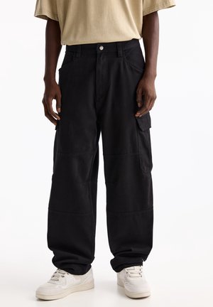 Dorosły ubrany w luźne czarne spodnie typu cargo, beżowy t-shirt i białe trampki, stojący na tle jednolitego białego tła.