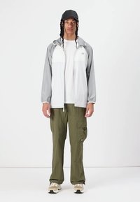 Jachetă ușoară gri și albă cu fermoar și glugă, combinată cu pantaloni cargo oliv și adidași bej. Include o șapcă neagră și un tricou alb.
