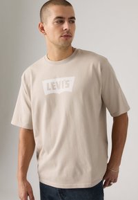 Beige bomulls t-skjorte med kort erm og rund hals, med en hvit Levi's logo på brystet. Ledig passform og myk tekstur.