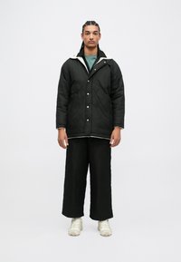 Giubbotto imbottito nero con colletto in pile bianco, abbinato a pantaloni neri larghi e sneaker chiari. Design minimalista, senza fantasie.