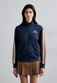 Navy blue zip-up jacket in smooth fabric, featuring white embroidery, with brown and white striped accents on sleeves. Paired with a pleated brown skirt.