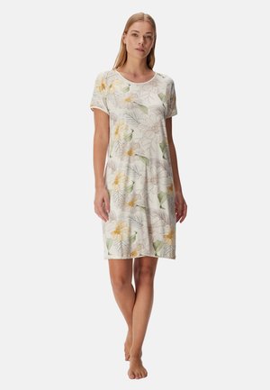 Femme debout pieds nus portant une robe à manches courtes, longueur genou, avec un motif floral pâle sur un fond clair.