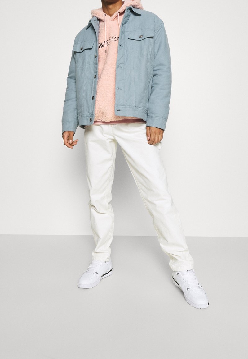 Lichtblauwe denimjacket over een roze fleeceshirt, gecombineerd met witte broek en witte sneakers. De outfit heeft een ontspannen pasvorm en contrasterende kleuren.