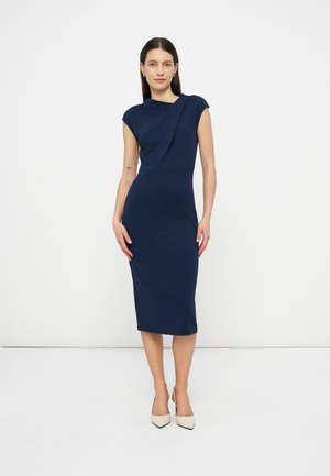 Frau trägt ein ärmelloses, marineblaues Midikleid mit asymmetrischem Ausschnitt und weißen spitzen Pumps, steht vor einem einfarbigen weißen Hintergrund.