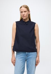 Top senza maniche navy con colletto, realizzato in tessuto liscio. Presenta una vestibilità rilassata e sottili spacchi laterali. Abbinato a denim azzurro chiaro.