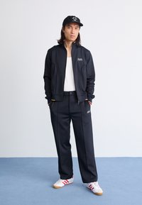 Jacket atlético preta com logótipo, top branco, calças pretas com logótipo, e sapatilhas brancas com riscas vermelhas em chão azul. Boné com design gráfico.