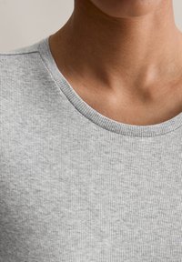 Közeli felvétel egy személy nyakáról és felső mellkasáról, amint világosszürke bordázott kereknyakú pólót visel, semleges háttér előtt.