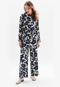 Blommig jumpsuit i mörkblått och krämfärgat, tillverkad av lättviktsmaterial. Har lång ärm, krage och vida byxben.