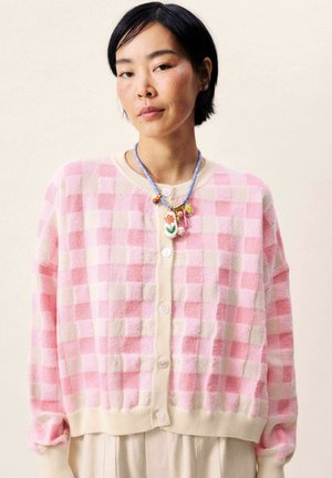 Jeune femme portant un cardigan à carreaux rose et blanc et un collier coloré en perles avec des pendentifs floraux et des breloques.