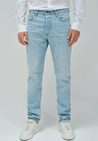 Jeans de mezclilla azul claro con un corte clásico, diseño de cinco bolsillos y botón marrón en la cintura. Combinados con zapatillas blancas.