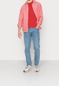 Rød kortærmet t-shirt under en lys rød skjorte med knapper; lys blå jeans; grå sneakers med brune accenter. Afslappet outfit, set forfra.