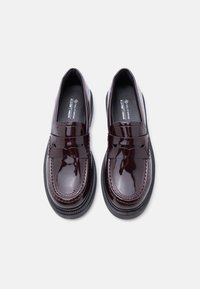 Mocassins en cuir verni bordeaux avec une finition lisse, bout rond, semelle plate, et une lanière décorative traversant le cou-de-pied. Matériaux véganes utilisés.