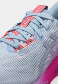 Αθλητικό παπούτσι ανοιχτού γαλάζιου χρώματος με λευκά κορδόνια, ετικέτα γλώσσας σε κοραλί ροζ και μωβ λογότυπο σε υφή στην επάνω επιφάνεια, τοποθετημένο σε ροζ σόλα.