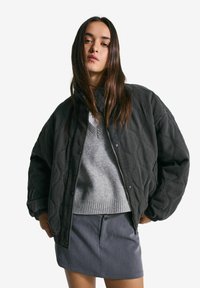 Mlada ženska z dolgimi rjavimi lasmi, oblečena v črno prešito jakno, sivo pulover in sivo krilo pred enobarvno belo ozadje.