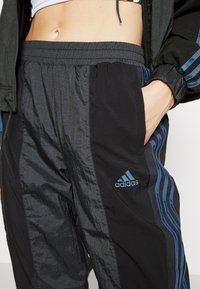 Gros plan d'une personne portant un pantalon de survêtement Adidas noir avec des bandes bleues et un logo, main dans la poche, et un top court dévoilant un ventre nu.