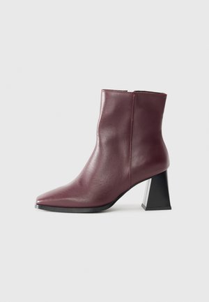 Stivale alla caviglia in pelle bordeaux con punta affilata, forma strutturata e tacco a blocco con accento nero. Texture liscia e cuciture minimali.