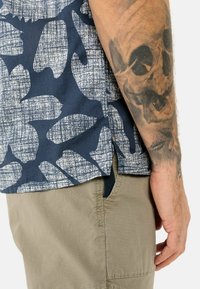 Kurzarmhemd in Marineblau mit einem weißen abstrakten Muster; aus einem strukturierten Stoff und mit einem gespaltenen Saum; kombiniert mit beigen Hosen.