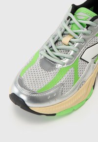 Scarpa atletica argento e verde con dettagli in mesh, superfici testurizzate e suola in gomma nera. Presenta accenti verdi e lacci beige.