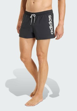 Hombre con bañador negro de Adidas con cordón blanco y gran logo blanco en el muslo izquierdo, de pie descalzo sobre un fondo claro.