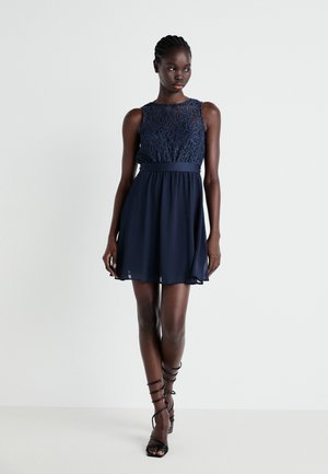Navy dress featuring a lace bodice, sleeveless design, flared skirt, and a fitted waistband. Worn with strappy black heels.
