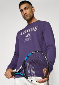 Lila kapucnis pulóver, rajta krémszínű "ADIDAS" és "TENNIS CLUB" felirattal. A modellen fekete és kék teniszütő van, amelynek mintás markolata van.