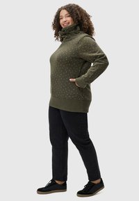 Sweatshirt vert olive avec col montant, motif à pois blancs, poignets et ourlet côtelés. Associé à un jean noir et des chaussures noires.