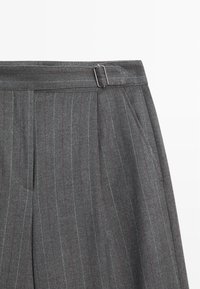 Pantalon gris à rayures fines avec devant plat, doté d'une boucle métallique et de plis, fabriqué dans un tissu texturé avec des variations subtiles de couleur.