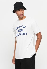 Hvit bomull T-skjorte med blå tekst "TRUCK FORD COUNTRY". Sammen med en svart bøttehatt, står modellen foran en enkel bakgrunn.