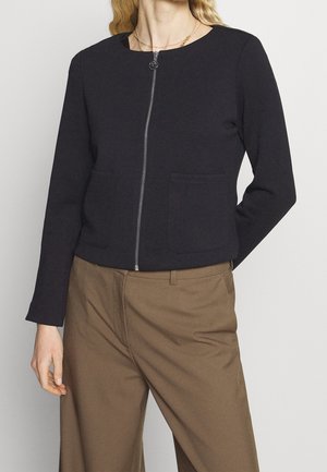 Femme portant une veste noire zippée avec deux poches avant et un pantalon marron à jambes larges avec passants pour ceinture.