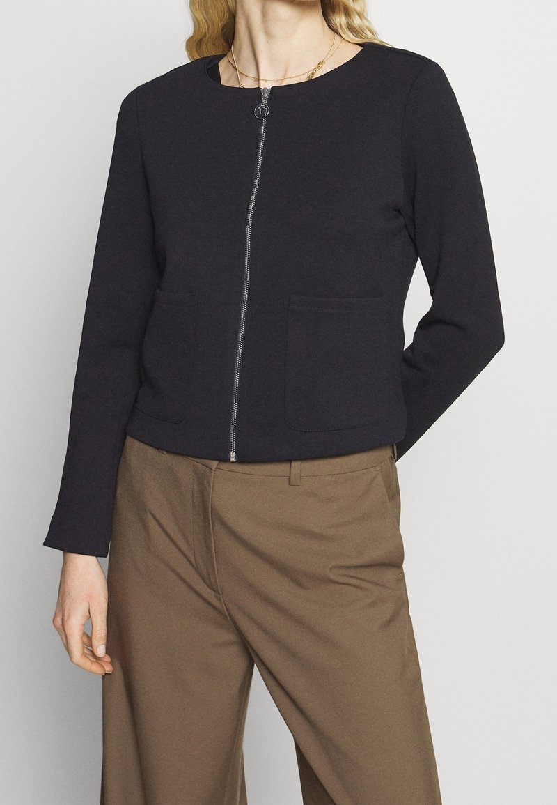 Femme portant une veste noire zippée avec deux poches avant et un pantalon marron à jambes larges avec passants pour ceinture.