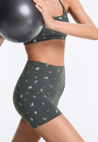 Femme portant un brassière de sport foncé à motifs floraux et un short taille haute, tenant un ballon d'exercice noir contre sa poitrine.