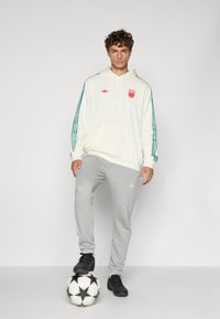 Cream hoodie with green stripes, red logo, and kangaroo pocket; gray sweatpants with white stripes and black soccer shoes on a ball.
