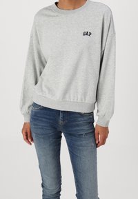 Sudadera de algodón gris con cuello redondo y mangas globo, con un pequeño logo negro de "GAP" en el pecho. Combinada con jeans azules.