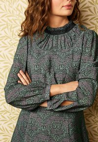 Vestido verde de manga larga con un patrón floral, escote fruncido y mangas acampanadas. La tela presenta tonos apagados de púrpura y hojas verdes.