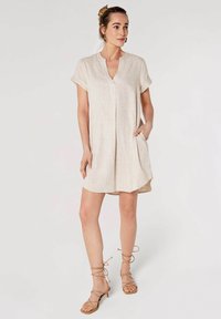 Robe en lin beige clair à manches courtes, décolleté en V et poches latérales. Le design présente une coupe décontractée et un ourlet légèrement arrondi.