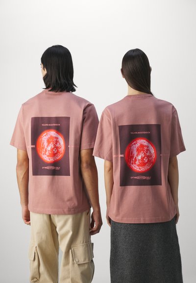 Pink cotton t-shirts with a rectangular graphic featuring a red planet. Text reads "YOU ARE AN EXTENSION OF THE UNIVERSE ITSELF."